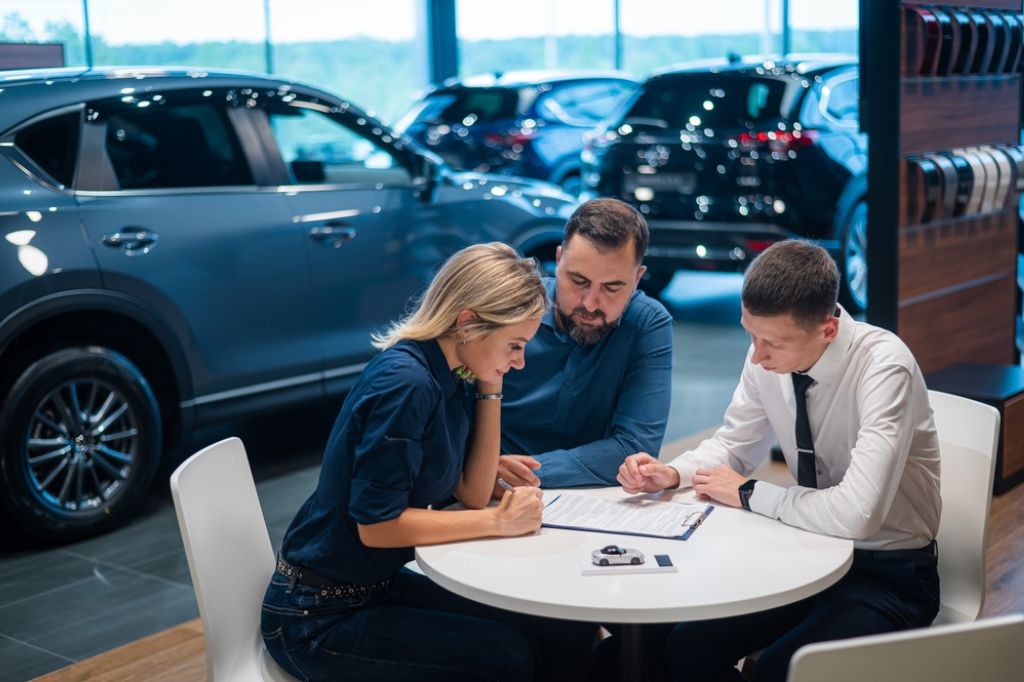 A couple makes use of the resurgent UK car market by signing a contract with a salesman. Cars can be seen in the background.